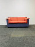 A modern two-seater sofa with orange cushions and blue-purple fabric arms and base, set against a plain white wall and gray carpet.