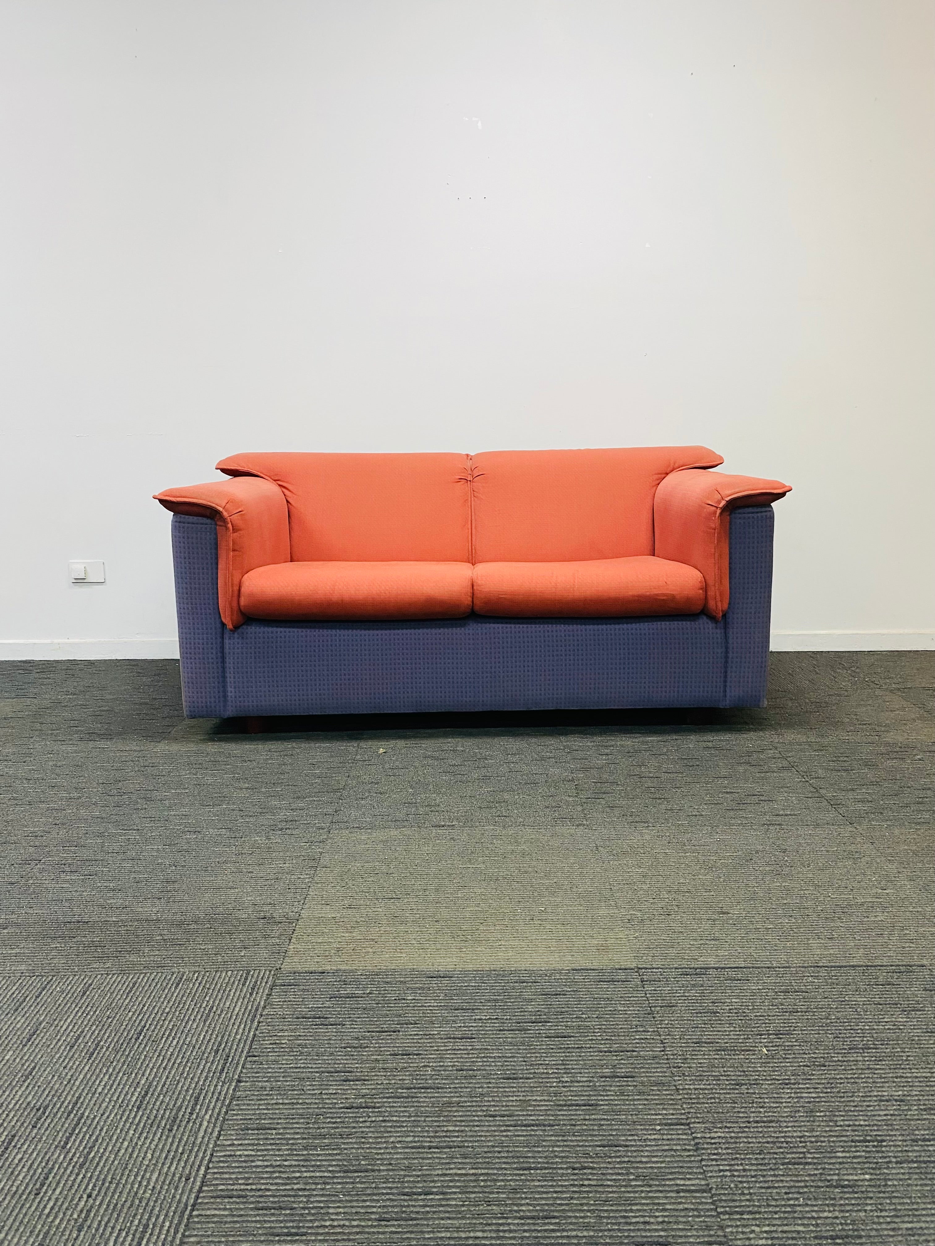A modern two-seater sofa with orange cushions and blue-purple fabric arms and base, set against a plain white wall and gray carpet.