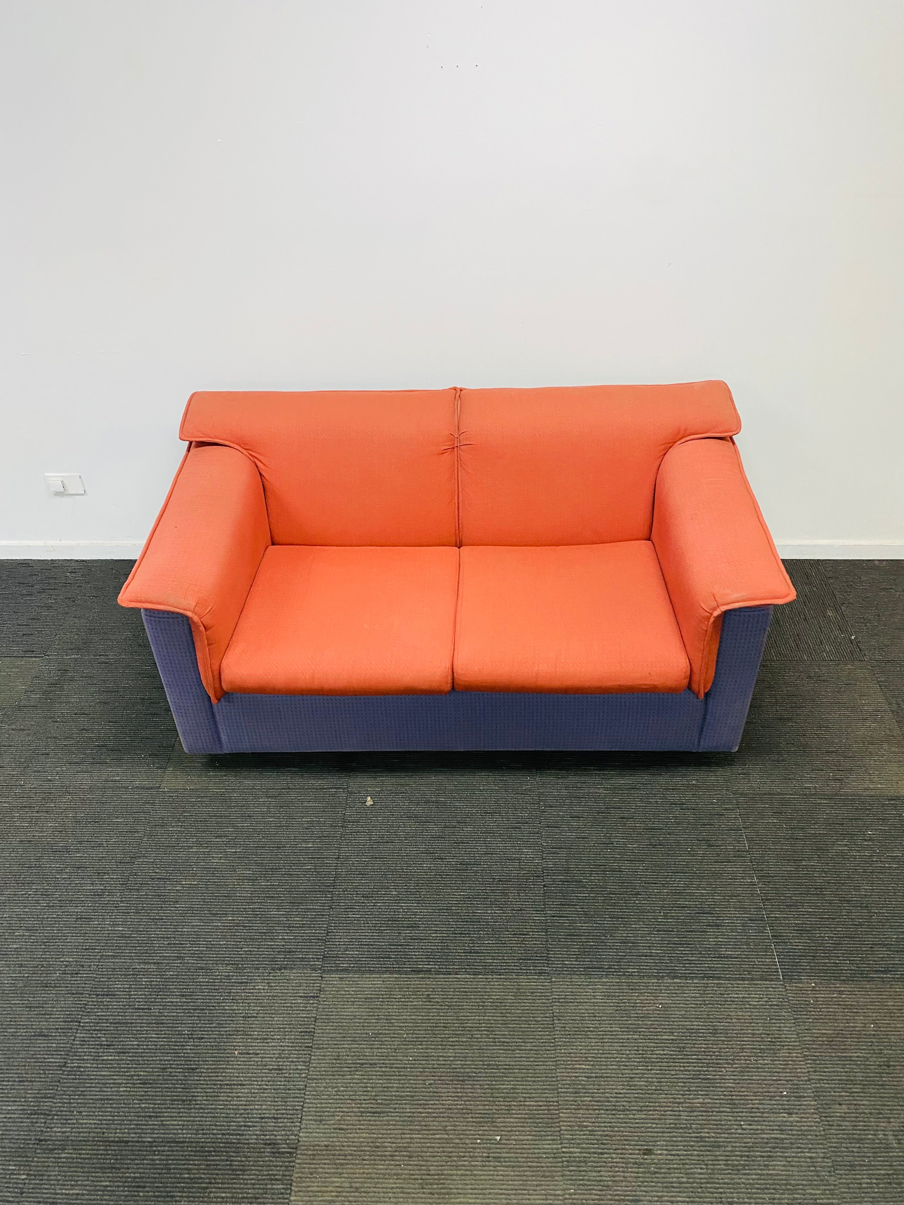 A two-seater loveseat sofa with orange upholstered seat and back cushions and a blue-purple base, photographed in an indoor room with a white wall and dark gray carpet.