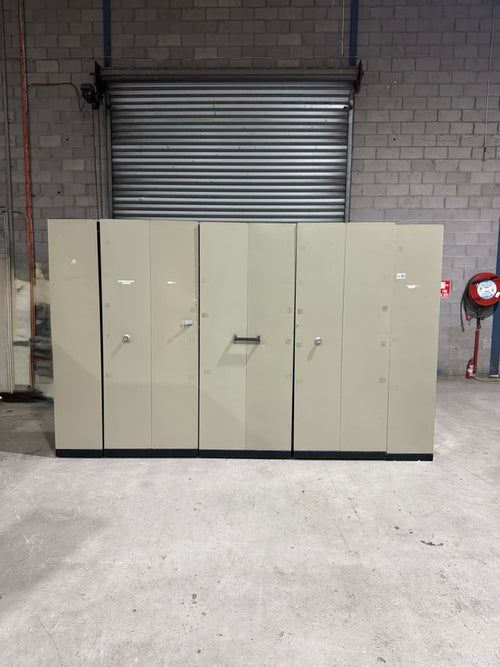 A set of five tall, light beige industrial metal storage cabinets with doors and handles, positioned in front of a gray brick wall and metal roll-up door.