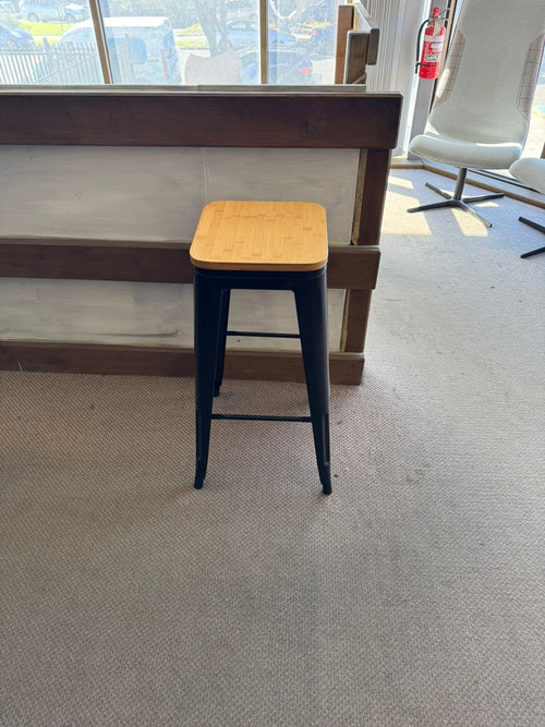 Bar stool with wooden seat and black metal legs in a room with large windows.