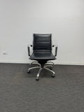 A black leather office chair with a five-star chrome base and armrests, positioned against a plain white wall on a dark gray carpeted floor.