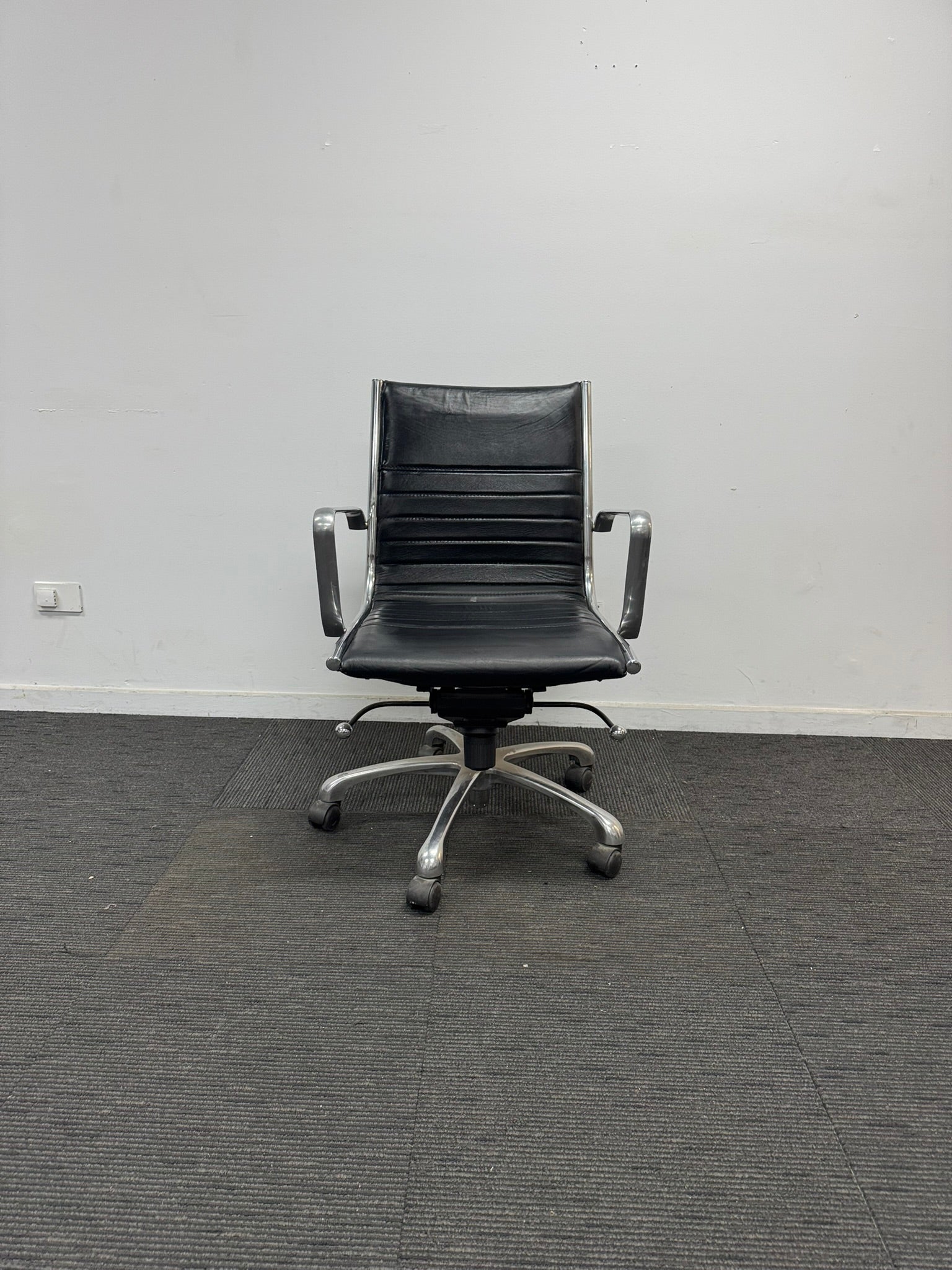 A black leather office chair with a five-star chrome base and armrests, positioned against a plain white wall on a dark gray carpeted floor.