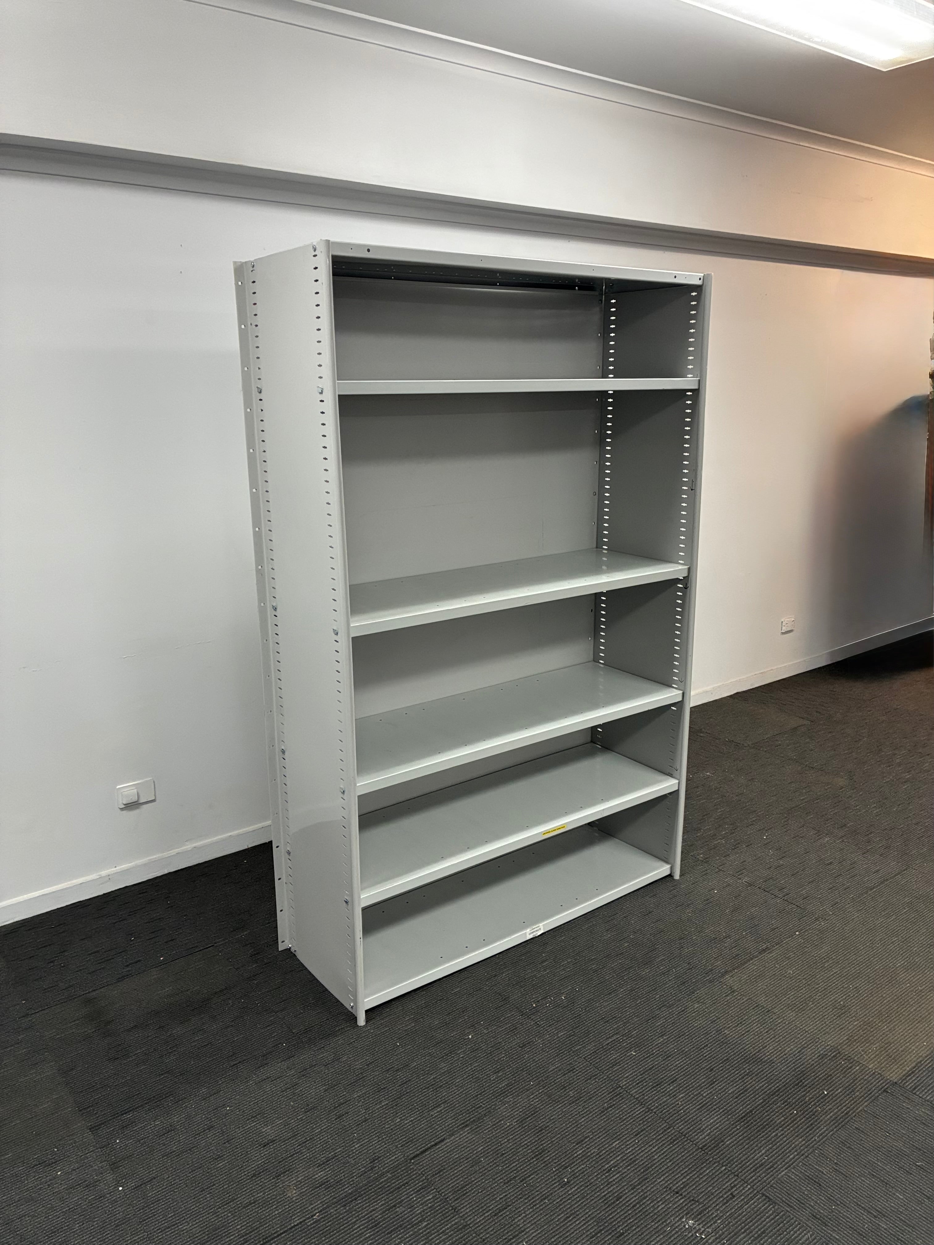 A tall, five-shelf gray metal bookcase standing in a room with white walls and dark gray carpet.