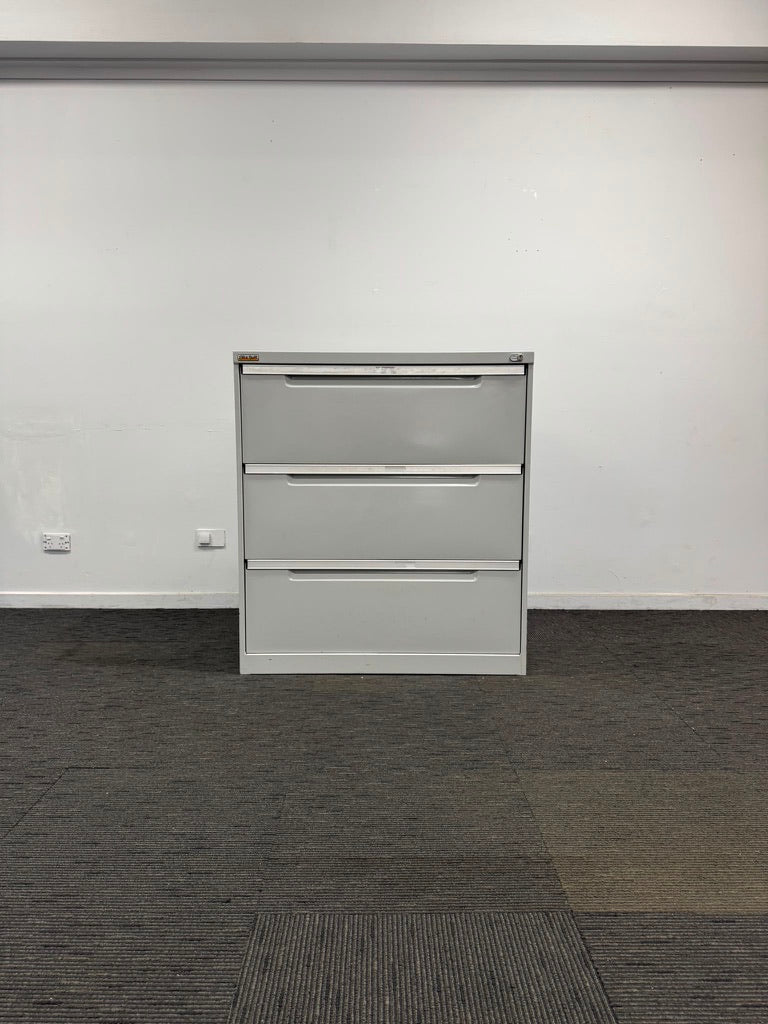 A three-drawer light gray metal file cabinet standing against a plain white wall on a dark gray carpeted floor.