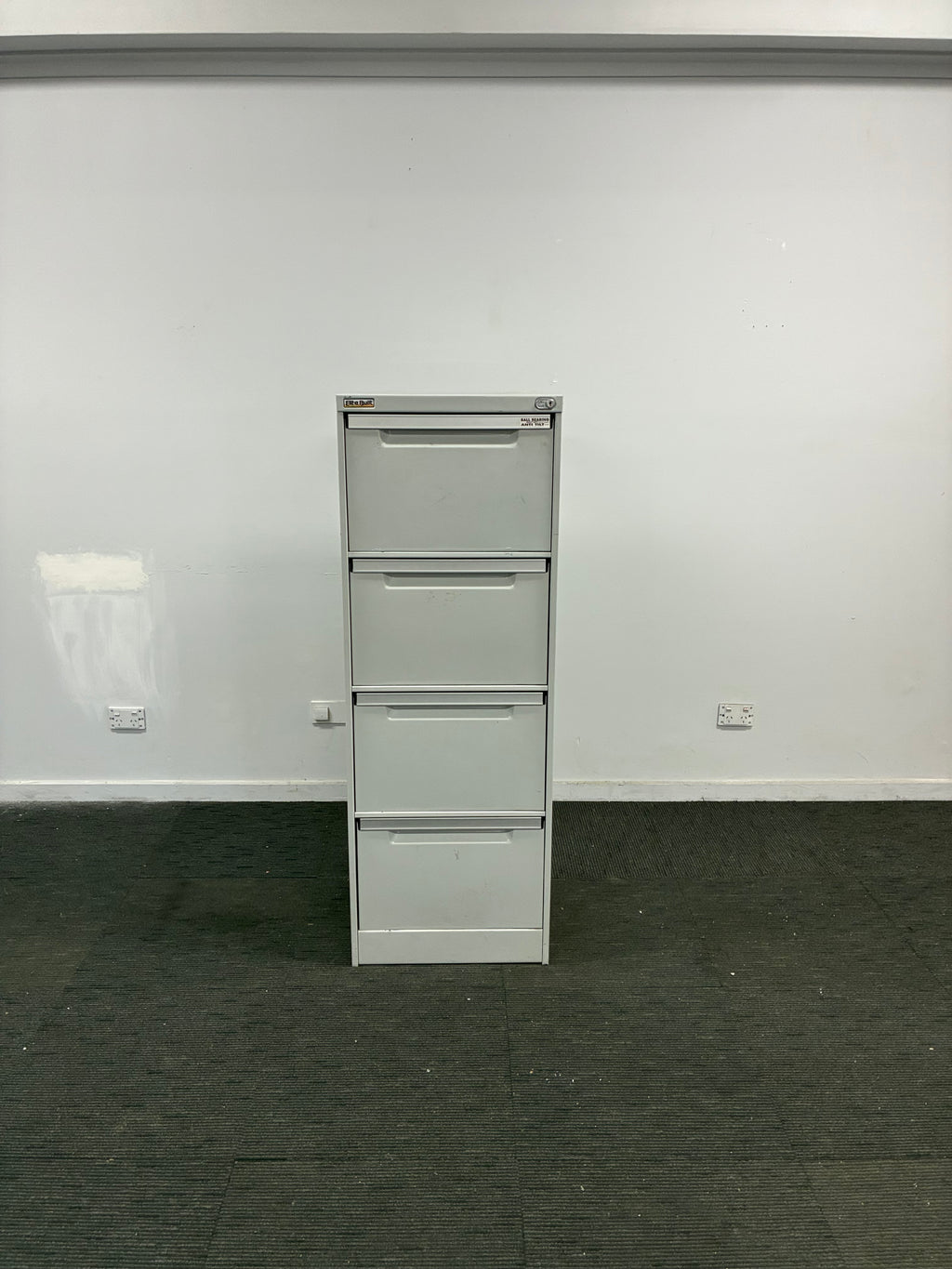 A four-drawer white metal file cabinet or storage unit mounted against a white wall, with a dark gray floor visible on the right side.