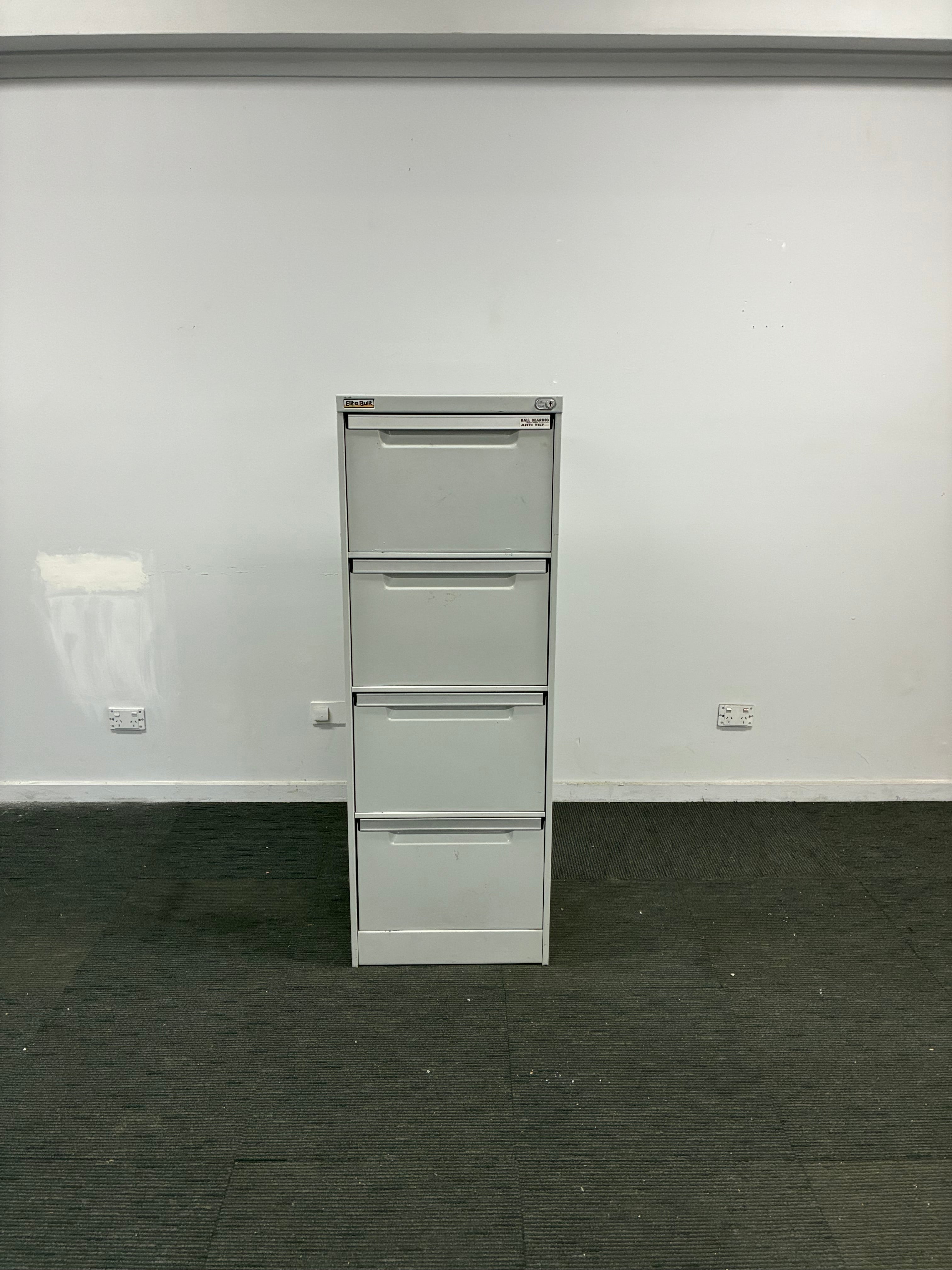 A four-drawer white metal file cabinet or storage unit mounted against a white wall, with a dark gray floor visible on the right side.