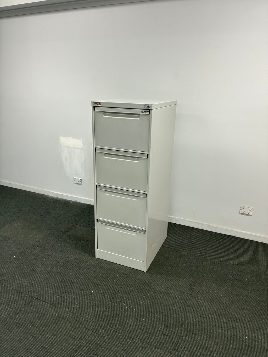 A white three-drawer office file cabinet with metal handles, shown in a room with a white wall and dark gray carpet.