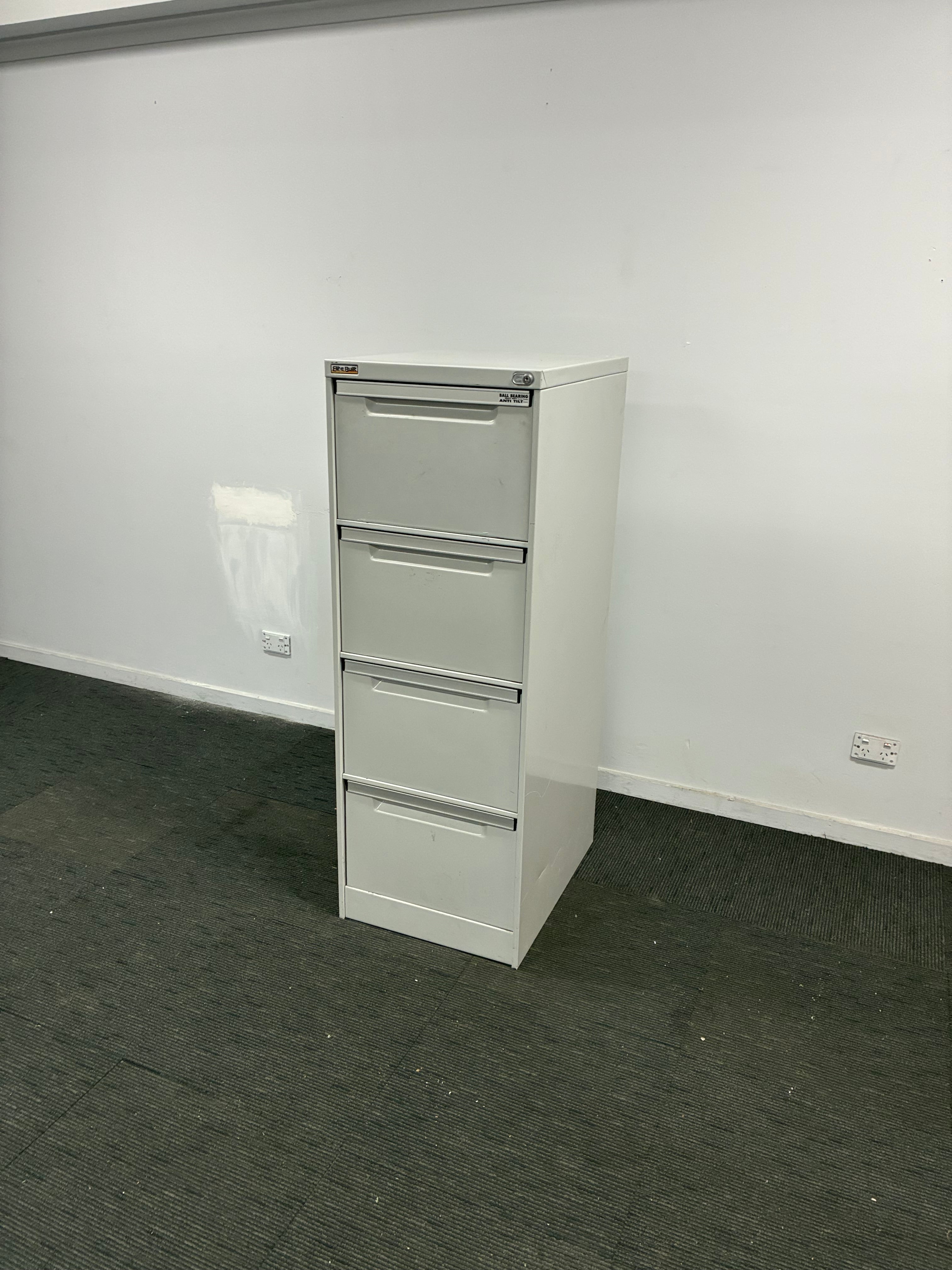 A white three-drawer office file cabinet with metal handles, shown in a room with a white wall and dark gray carpet.