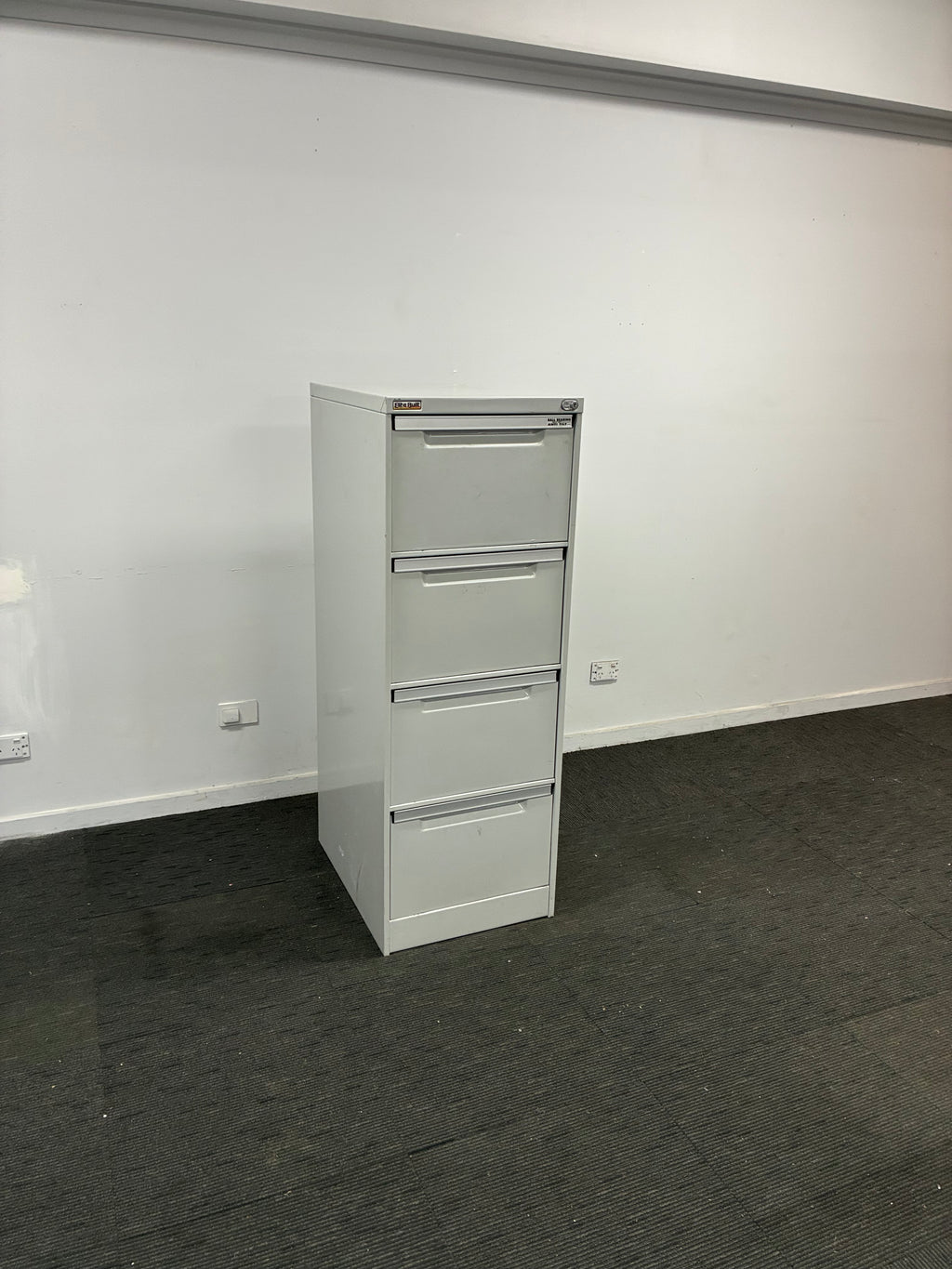 A white four-drawer metal file cabinet mounted on a white wall, shown in a room with a dark gray carpeted floor.
