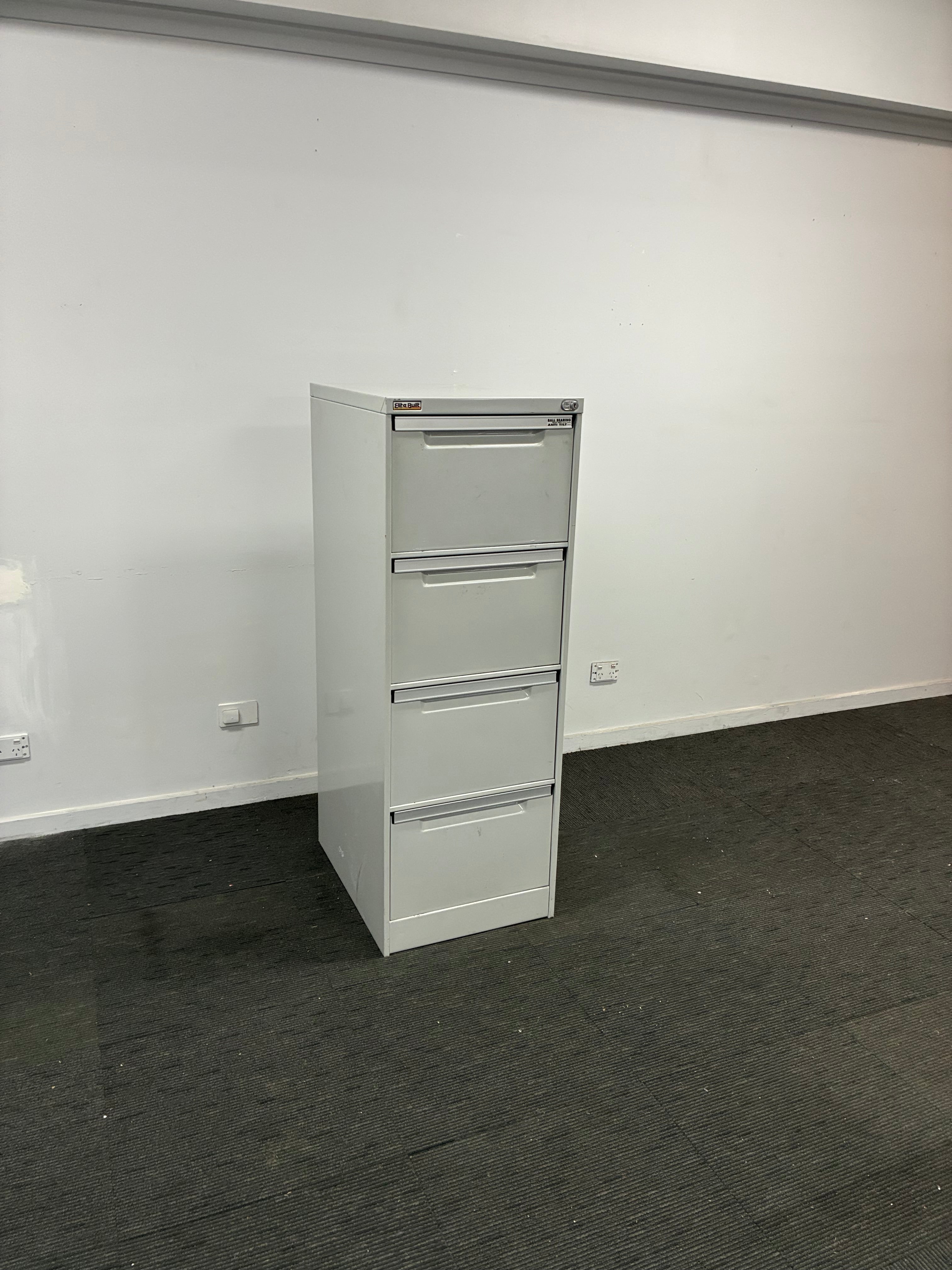A white four-drawer metal file cabinet mounted on a white wall, shown in a room with a dark gray carpeted floor.