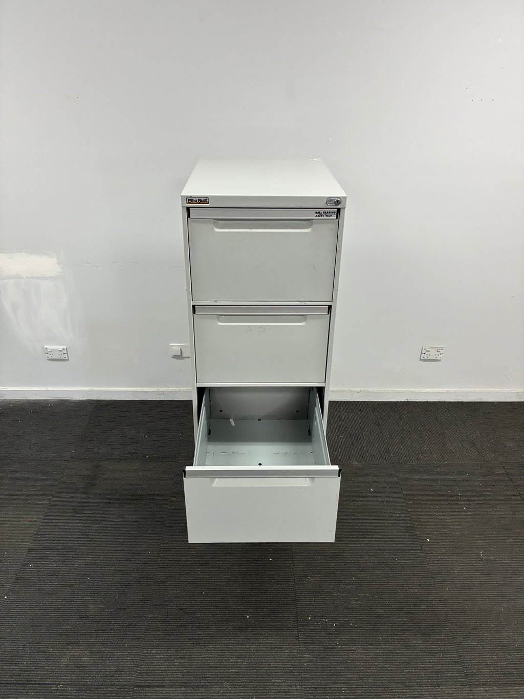 A white metal two-drawer filing cabinet with one drawer open, shown against a white wall and dark gray carpeted floor.