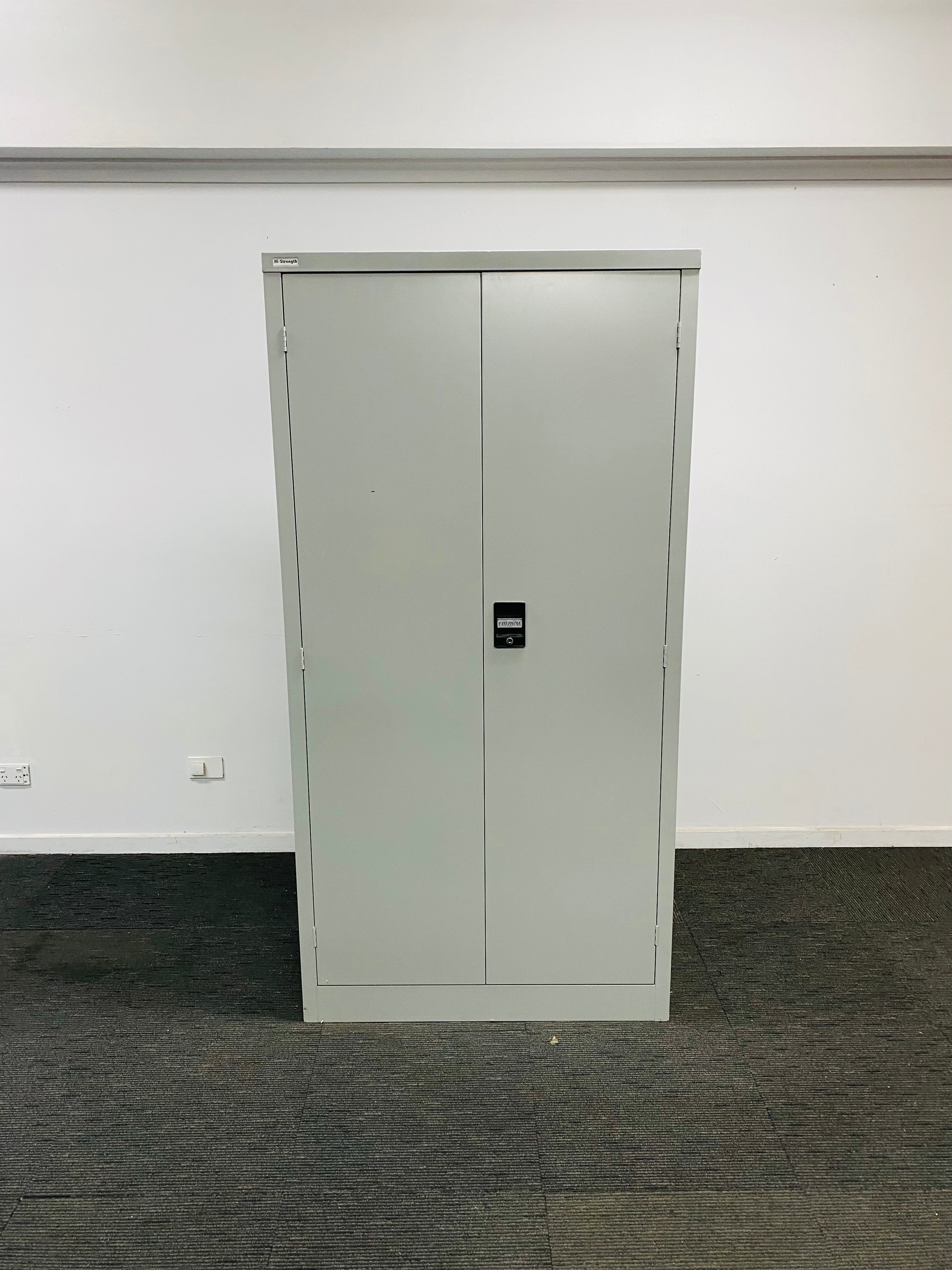 A tall, light gray metal double-door storage cabinet with a small black digital lock, set against a plain white wall and dark gray carpeted floor.