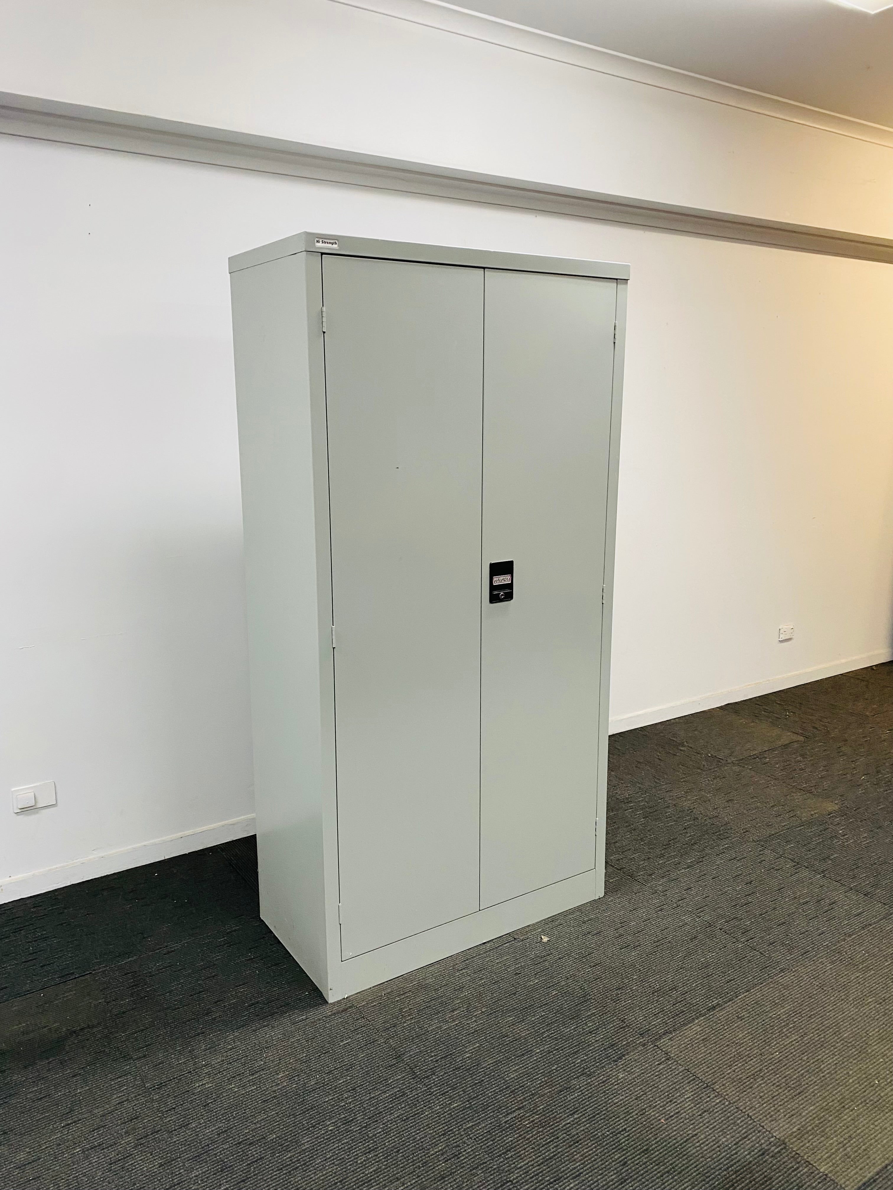 A tall, light gray metal two-door storage cabinet with a small black lock, standing on a dark gray carpeted floor against a white wall.