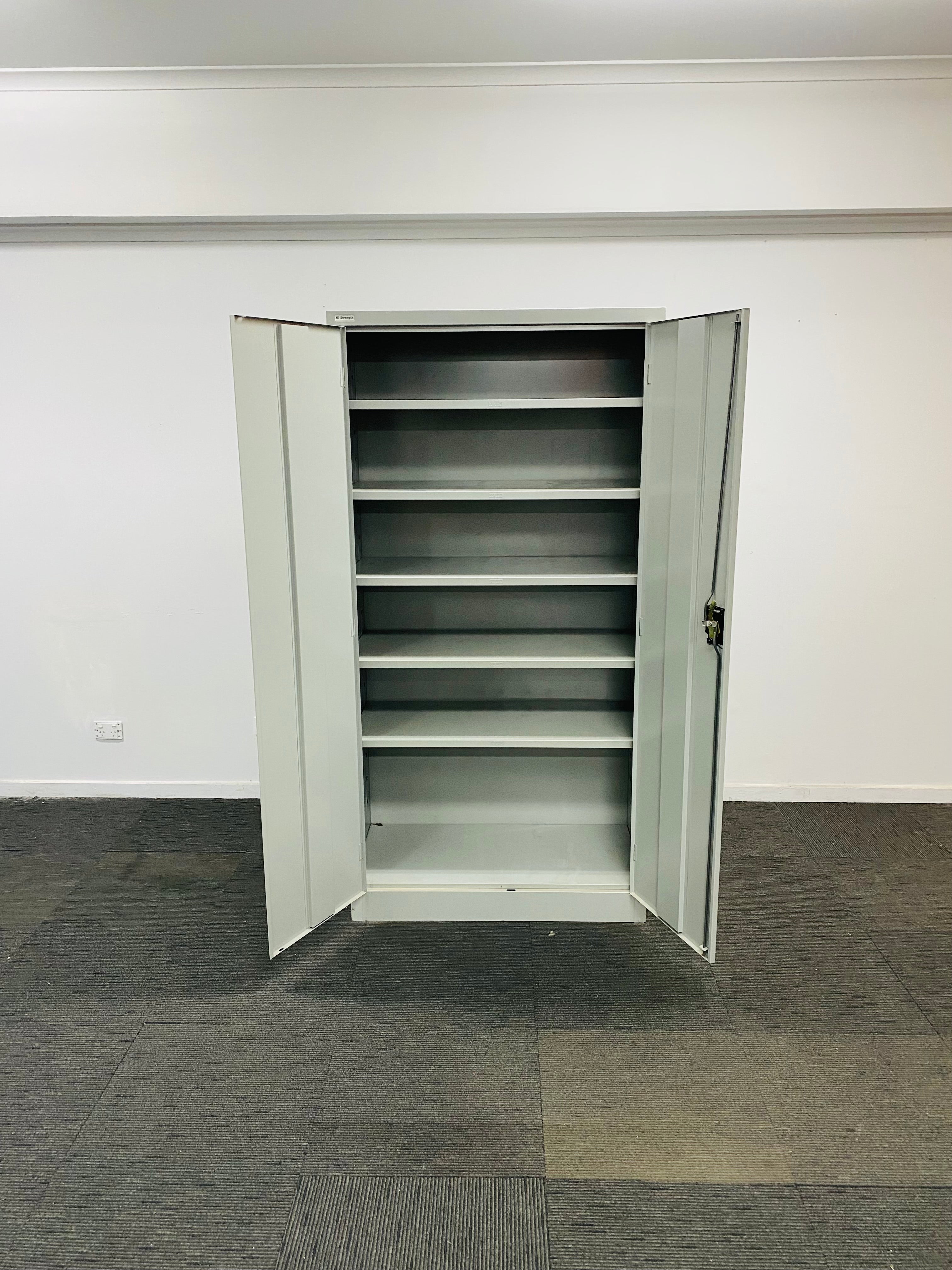 A tall, light gray metal storage cabinet with two doors open, showing five interior shelves, set against a plain white wall and gray carpeted floor.