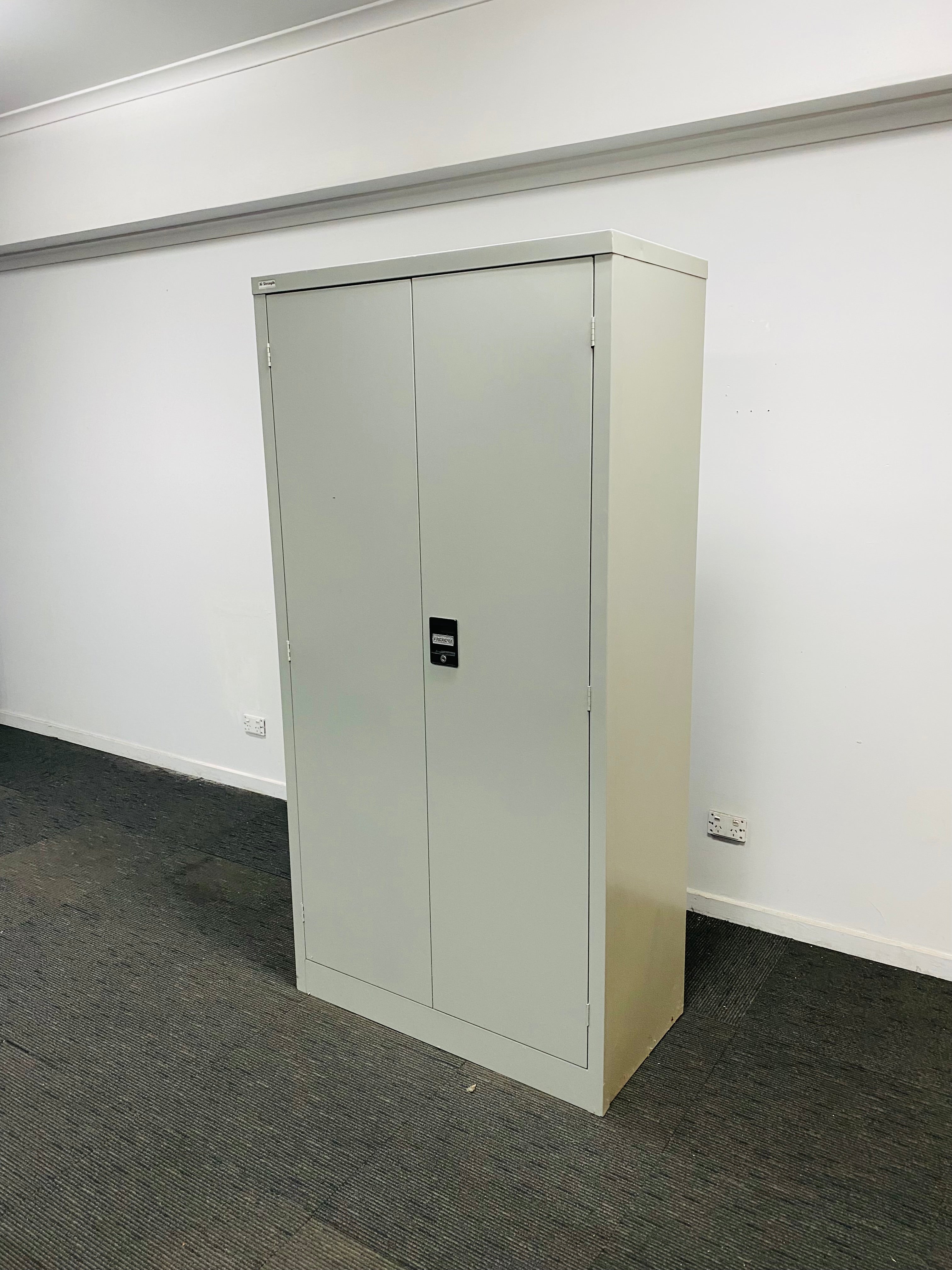 A tall, light gray metal two-door storage cabinet with a small black digital lock, standing on a dark gray carpeted floor against a white wall.