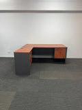 An L-shaped office desk with a reddish-brown wood-grain top and matching wood-grain side panels, paired with dark gray side bases, shown in an empty room with gray carpet and white walls.