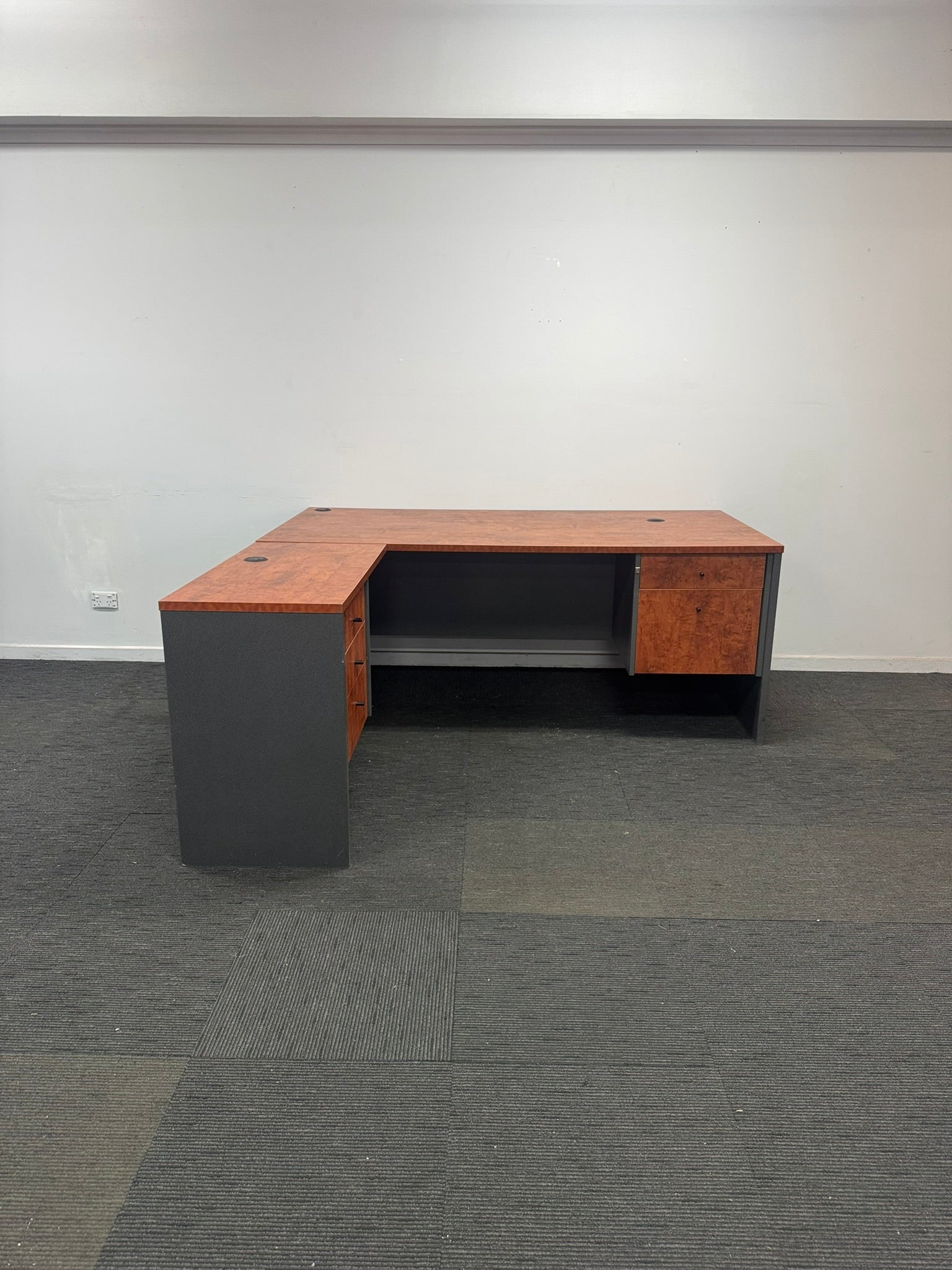 An L-shaped office desk with a reddish-brown wood-grain top and matching wood-grain side panels, paired with dark gray side bases, shown in an empty room with gray carpet and white walls.