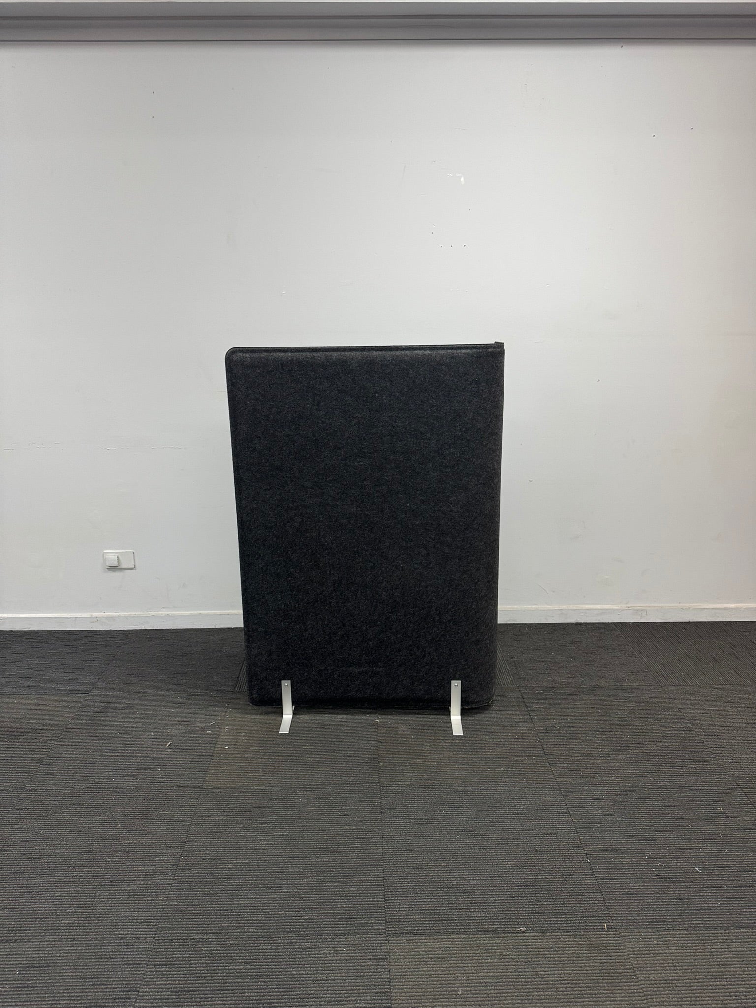 A tall, black upholstered office partition panel or speaker cabinet on small white support feet, set against a plain white wall and gray carpet.