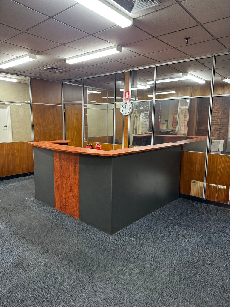 An L-shaped reception desk in a modern office setting, featuring a dark gray body, a warm wood-grain front panel, and a light wood countertop. The desk is situated in a space with gray carpet, glass partition walls, wood paneling, and a tiled ceiling with fluorescent lights.