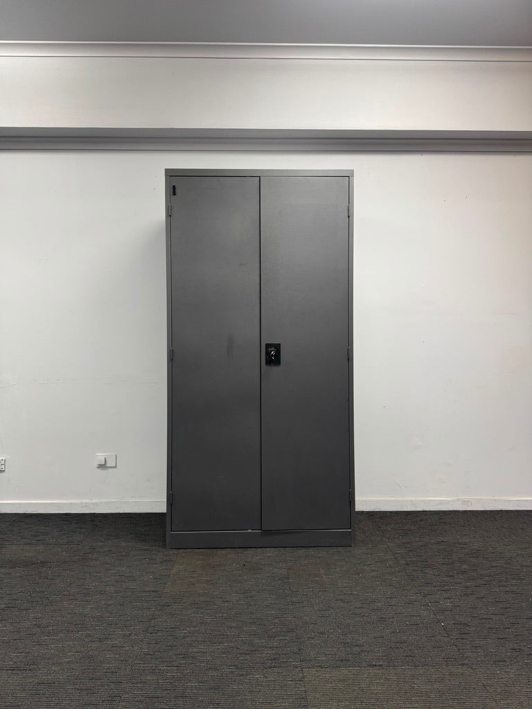 A tall, dark gray double-door metal cabinet or storage unit with a small lock, set against a plain white wall with a gray carpeted floor.