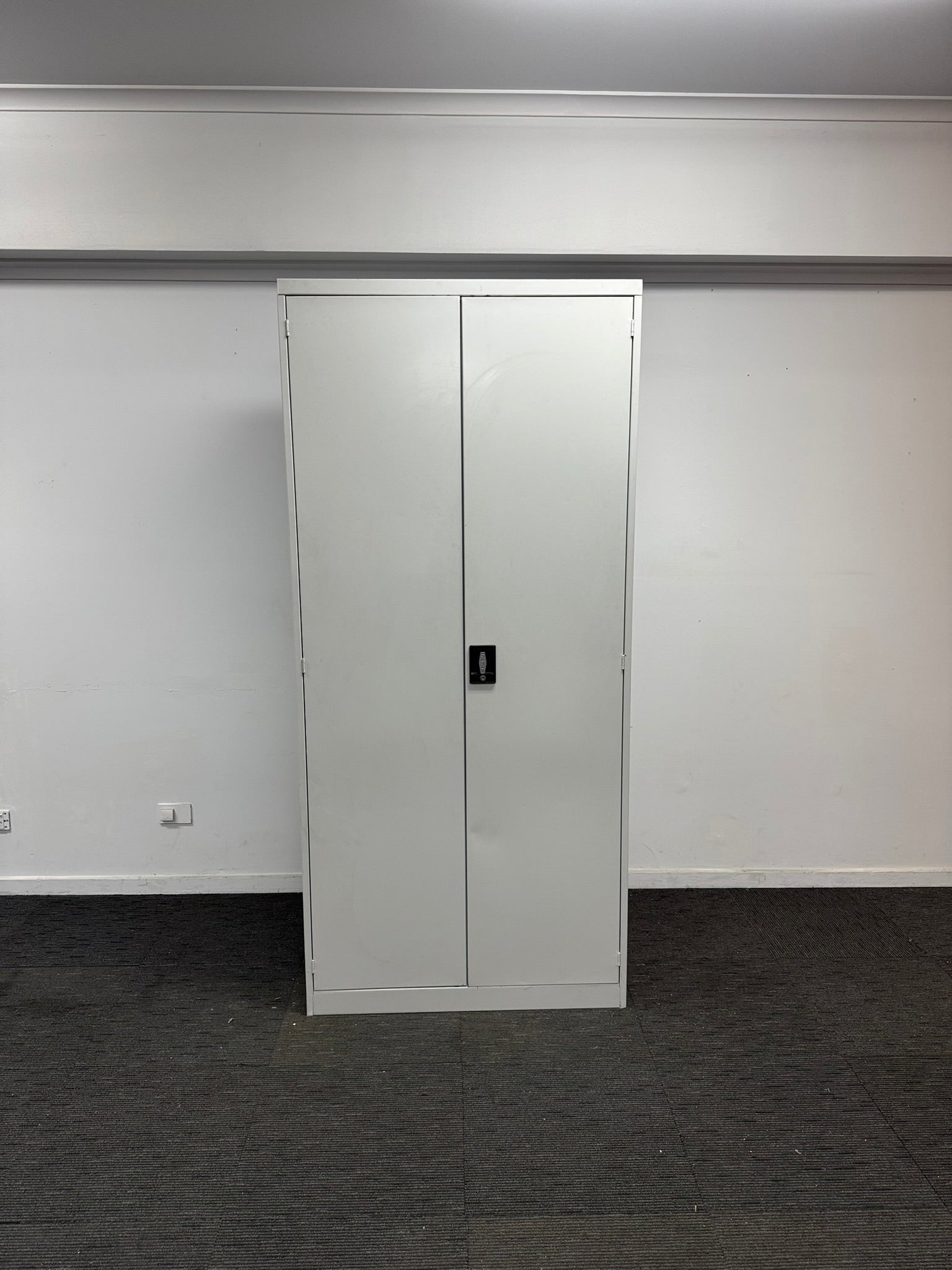 A tall, narrow white metal storage cabinet with two doors and a small black handle, set against a plain white wall with a dark gray carpeted floor.