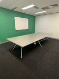 A long rectangular meeting table with a light gray top and slim metal legs, shown in a conference room with a green wall, whiteboard, dark gray carpet, and ceiling tiles.
