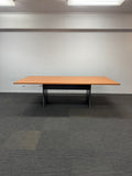 A rectangular meeting table with a light wood grain top and a black metal frame, set against a plain white wall and gray carpet.
