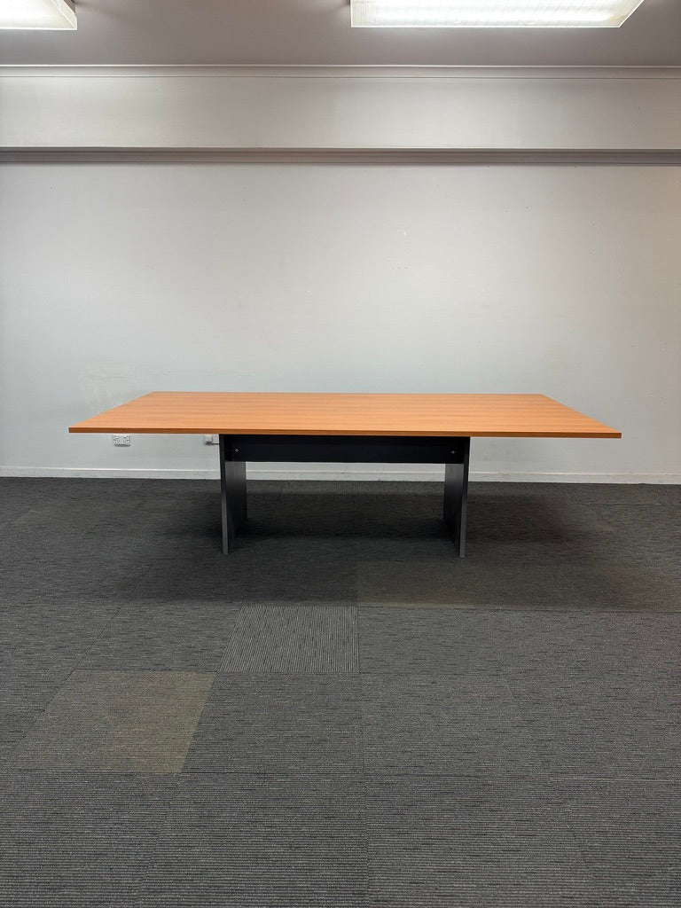 A rectangular meeting table with a light wood grain top and a black metal frame, set against a plain white wall and gray carpet.