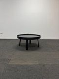 A black round coffee table with three legs, shown in a minimalist room setting with a white wall and gray textured carpet.