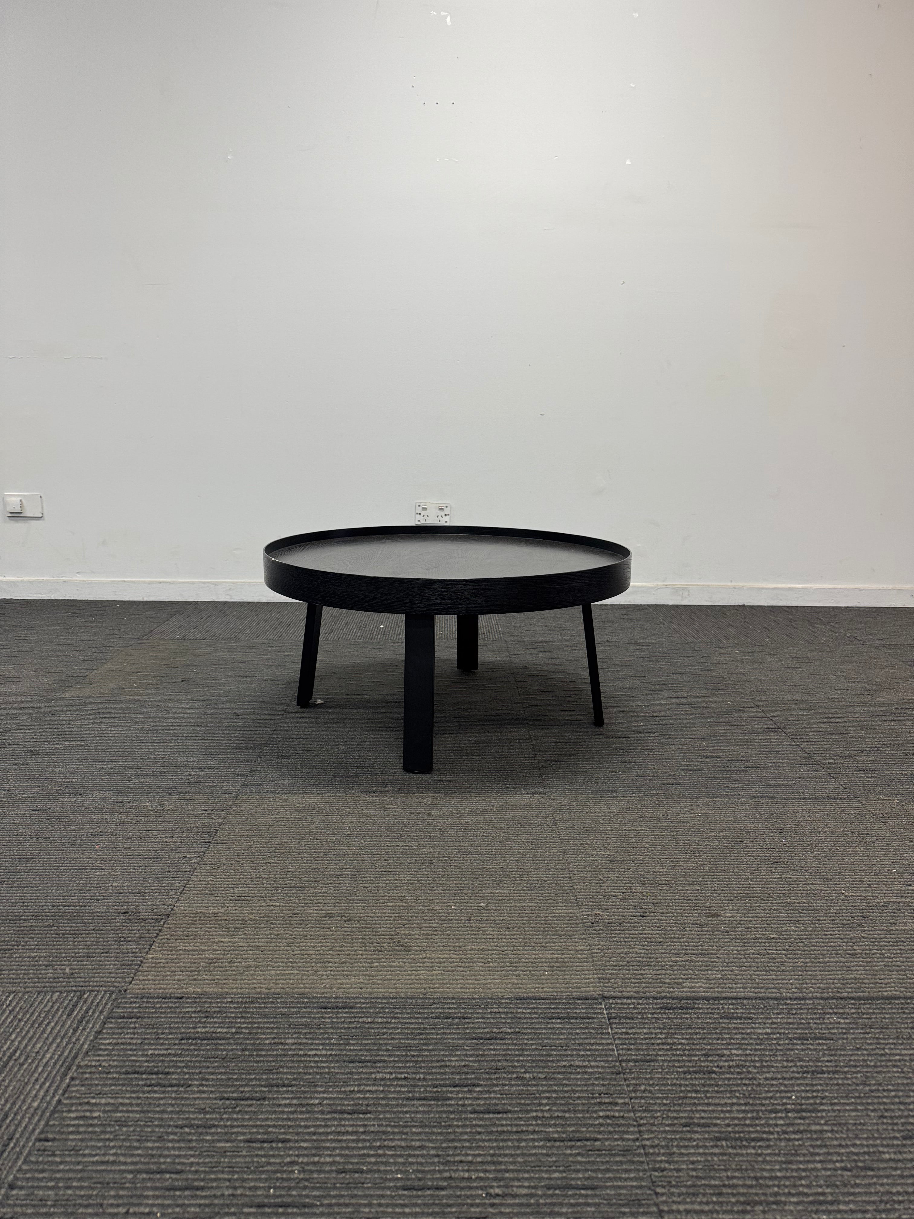 A black round coffee table with three legs, shown in a minimalist room setting with a white wall and gray textured carpet.