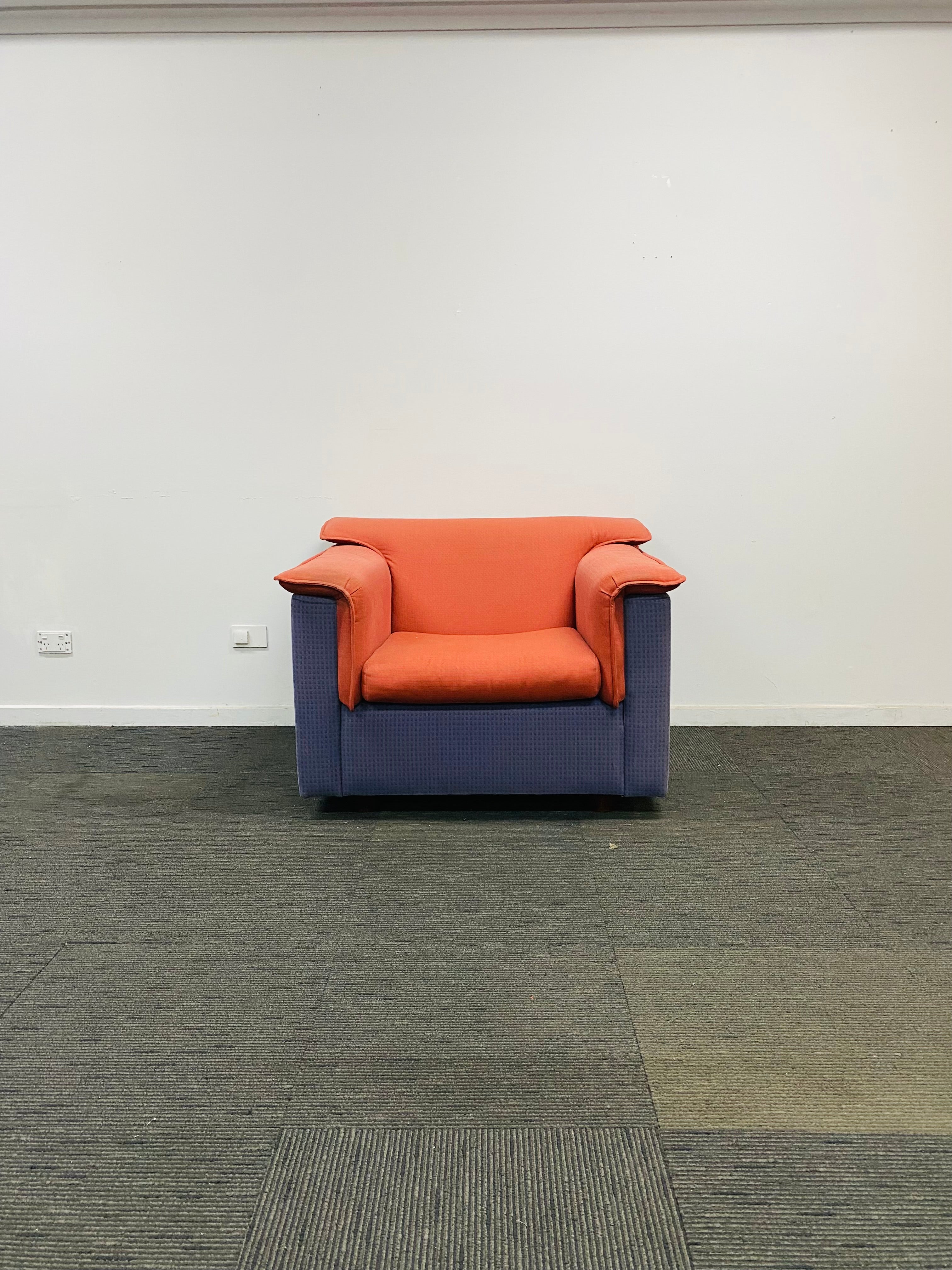 A modern accent armchair with a terracotta orange seat and back cushion paired with a slate blue-purple frame, set against a plain white wall and gray carpet.