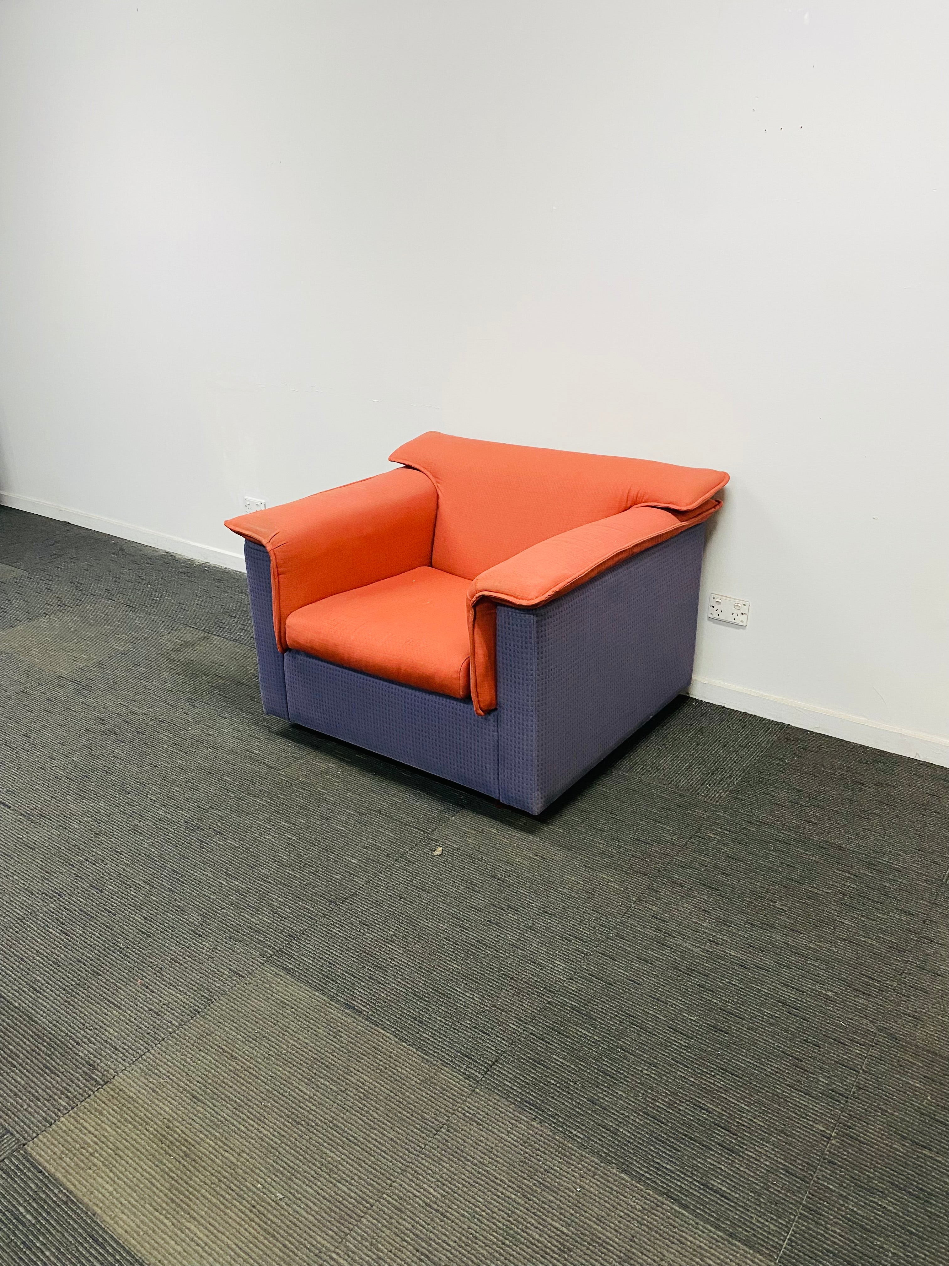 A modern armchair with orange seat and back cushions and a blue-purple upholstered base, placed on a gray carpeted floor against a plain white wall.