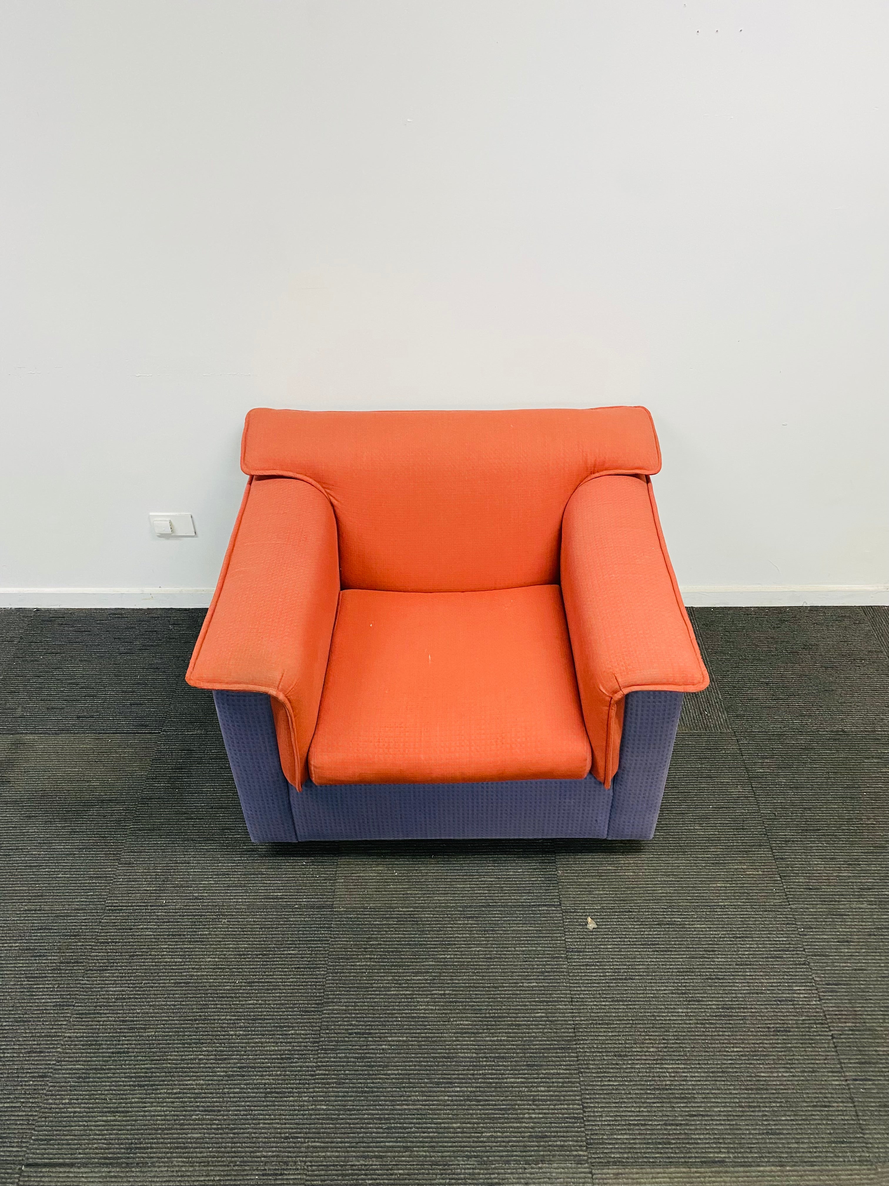 A modern accent chair with a bright orange upholstered seat and backrest, and a blue-purple ribbed fabric base, shown on a dark gray carpet against a white wall.