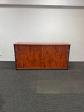 A mid-century modern wooden credenza or sideboard cabinet with two doors and a rich reddish-brown wood grain finish, set against a plain white wall and dark gray carpet.