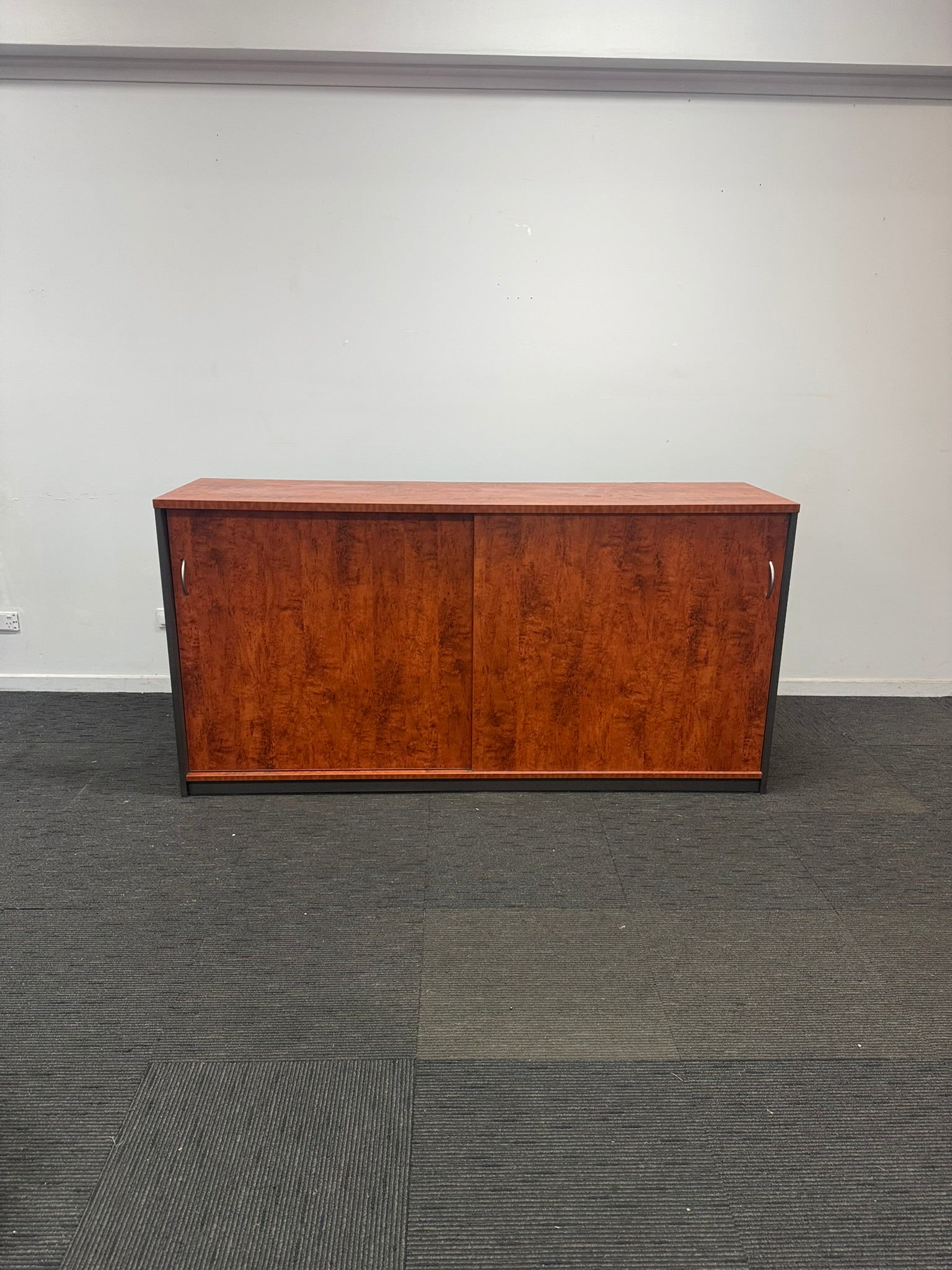 A mid-century modern wooden credenza or sideboard cabinet with two doors and a rich reddish-brown wood grain finish, set against a plain white wall and dark gray carpet.