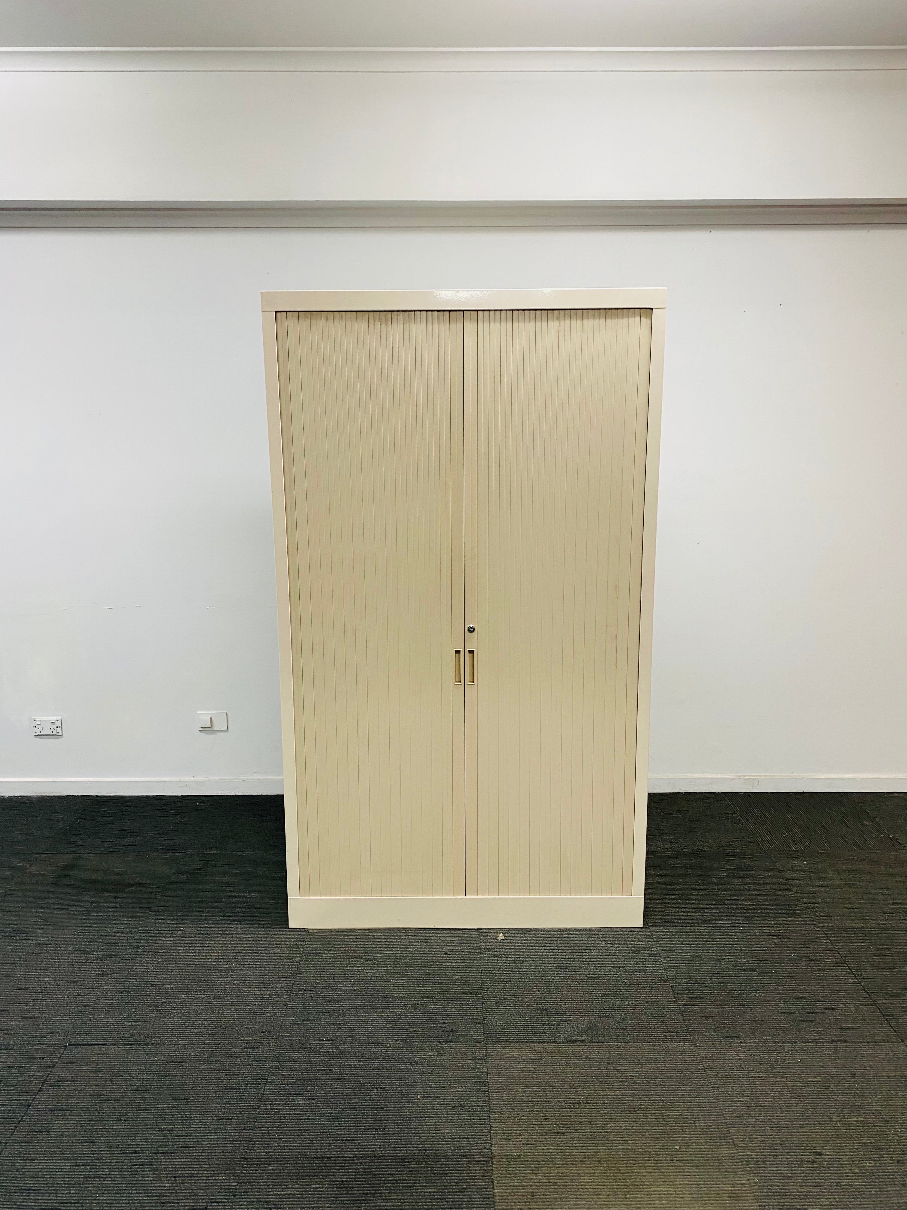 A tall, narrow double-door cabinet or wardrobe in a light beige woodgrain finish, standing on a dark gray carpeted floor against a plain white wall.