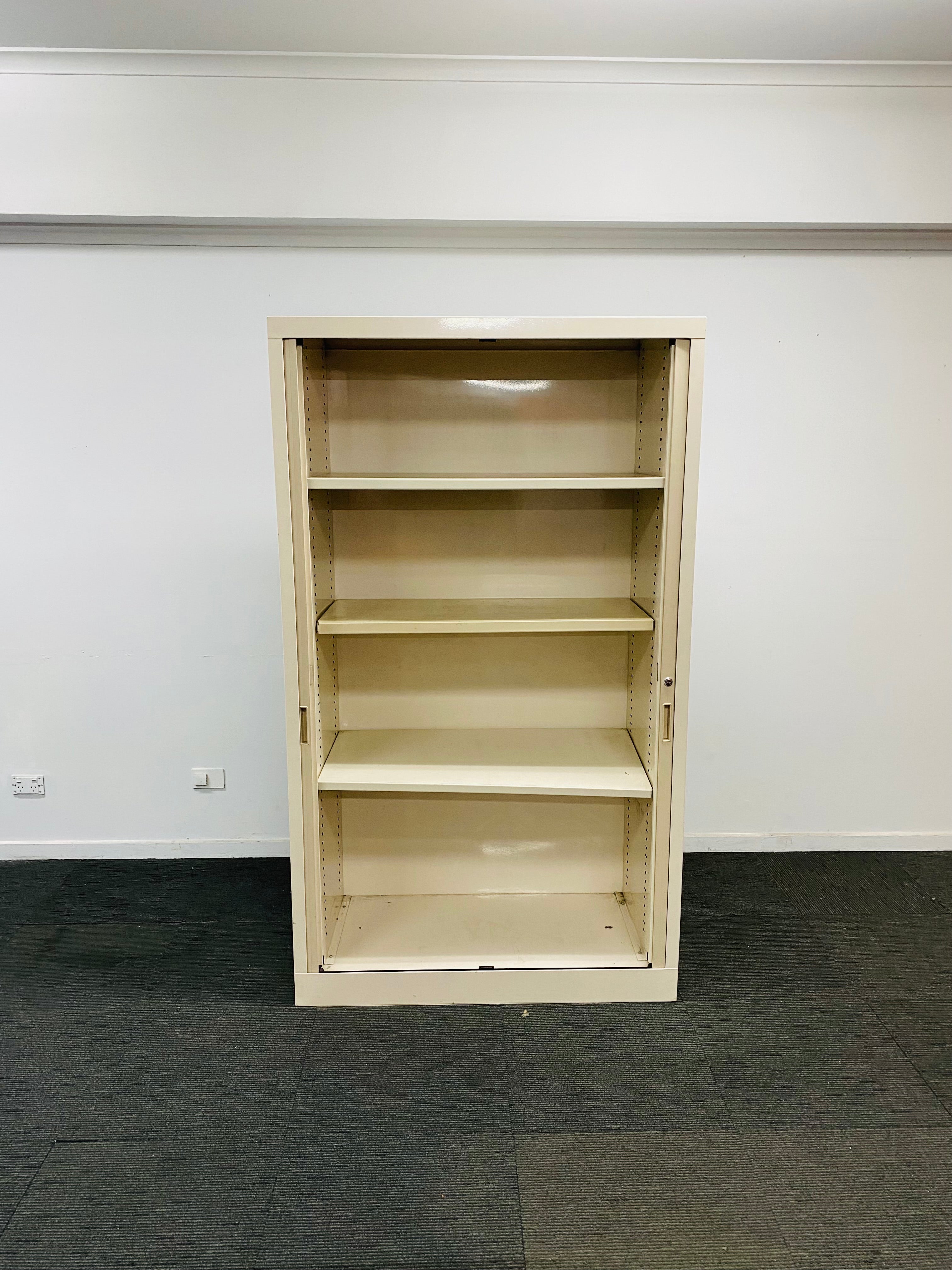 A tall, empty beige metal bookcase with four shelves, set against a plain white wall and dark gray carpet.