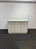 A white three-door sideboard cabinet with slim silver handles, set against a plain white wall and dark gray carpeted floor.