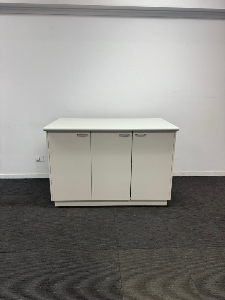 A white three-door sideboard cabinet with slim silver handles, set against a plain white wall and dark gray carpeted floor.