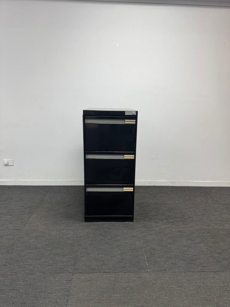 A black three-drawer metal file cabinet with silver handles, standing on a gray carpeted floor against a plain white wall.