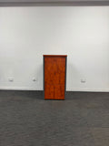 A tall, narrow wooden cabinet or storage unit with a reddish-brown burl wood finish and a dark gray side frame, standing on a dark gray carpeted floor against a plain white wall.