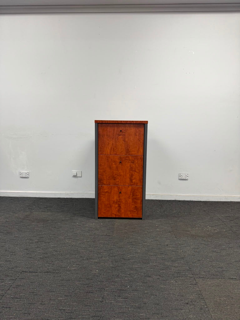A tall, narrow wooden cabinet or storage unit with a reddish-brown burl wood finish and a dark gray side frame, standing on a dark gray carpeted floor against a plain white wall.