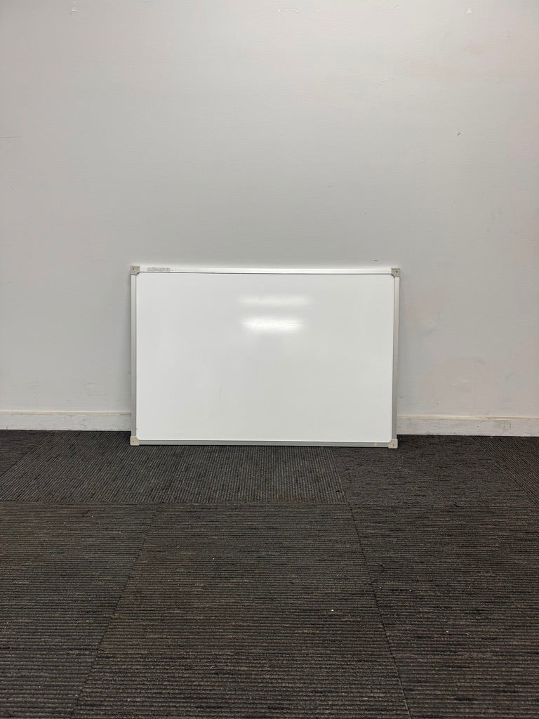 A white rectangular dry-erase whiteboard with a silver metal frame, leaning against a plain light gray wall on a dark gray carpeted floor.